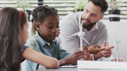Diverse male teacher and happy schoolchildren studying wind turbines in school classroom