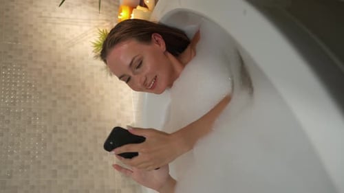 Woman Relaxes in Bubble Bath Looking at Phone