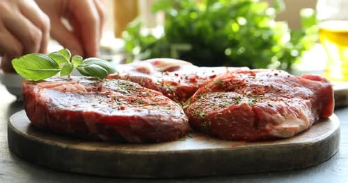 Woman sprinkling spices onto raw steaks at grey table, closeup