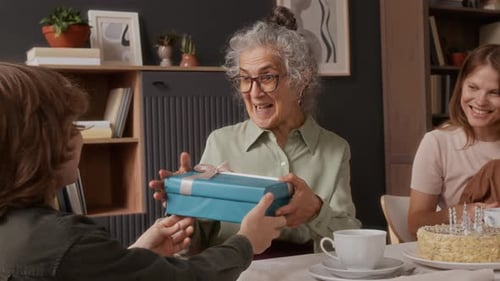 Niño sonriente regalando una caja de regalo a su abuela sorprendida durante la celebración de su cumpleaños
