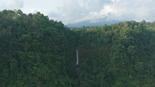 Aerial shot slowly approaching a tall, powerful waterfall flowing through a lush green jungle