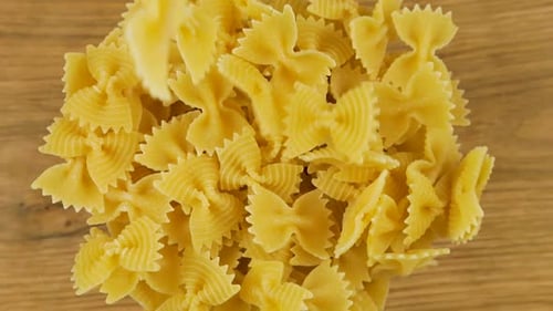 Bow-shaped pasta is poured into a glass transparent bowl against an oak wooden board. Top view. Slow
