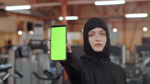 Athletic Muslim Woman Wearing Hijab and Sportswear and Shows Smartphone Green Screen Mockup in the