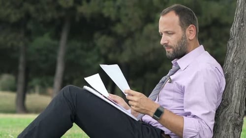 Young Businessman Working with Documents Sitting on Grass in Park 30s