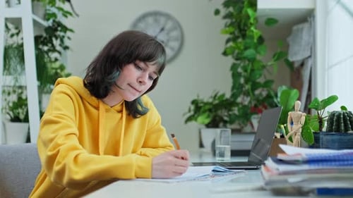 Teenage College Student at Home Using Laptop Writing in Notebook