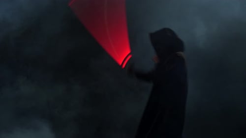 Young Man in Jedi Cosplay Costume with Lightsaber Battle on Black Background in Smoke and Rain Slow