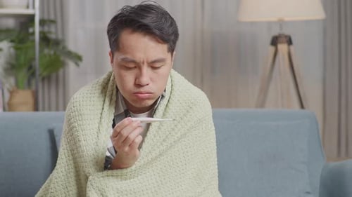 Close Up Of Asian Man With Blanket Using Thermometer Test For Flu, Cold And Body Temperature On Sofa