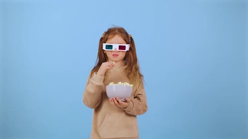 Child Wearing 3D Glasses Holds a Bowl of Popcorn Against a Blue Background While Enjoying a Movie