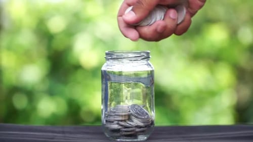 Human hand put and saving coins in a glass container with blur green nature background. Saving conce