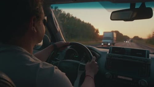 Young Man Steers the Car in Heavy Traffic on the Highway Full of Trucks