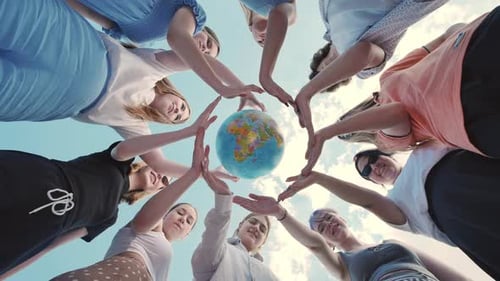 Gruppe junger Freunde, die mit ihren Händen einen Kreis um einen Globus vor blauem Himmel bilden