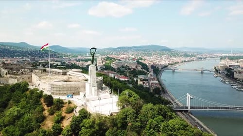 Liberty statue overlooking budapest cityscape with hungarian flag waving in citadel, dolly aerial