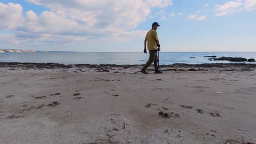 Man with Metal Detector Walking Along Sandy Beach Near the Sea Searching for Treasure Under Blue Sky