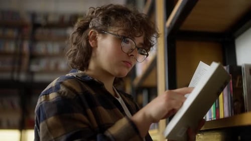 Confident Girl with Curly Hair Wearing Glasses Reads a Book Near the Shelves in a Library with a Lot