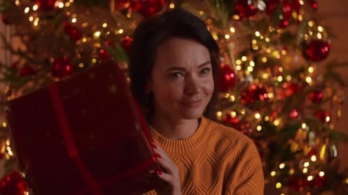 Woman Joyfully Shaking Gift by Christmas Tree
