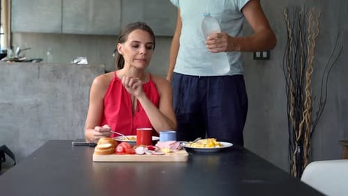 Woman Eating Breakfast While Man Pours Water at Home