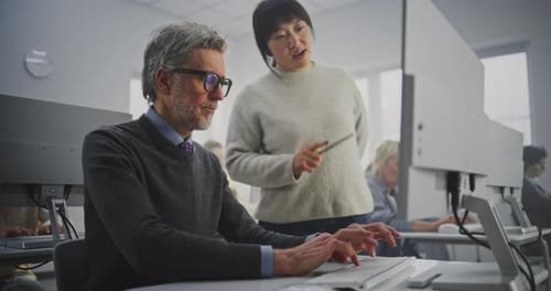 Elegant Grey Haired Man in Glasses Works on Computer While Young Teacher Leans Over Pointing Screen