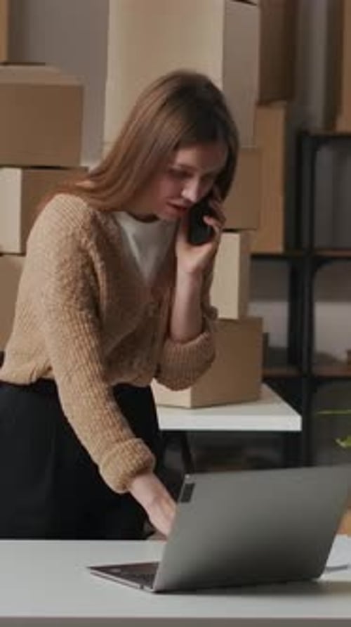Woman on Phone Working at Laptop with Boxes