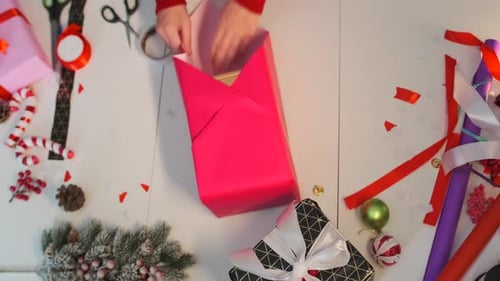 Top view of female hands wrapping Christmas gift with red paper and white ribbon.