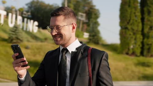 Outdoors Businessman in Suit with Glasses Stand Near Modern Building Holding Smartphone in Hands