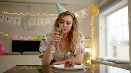 Woman in a Nice Dress and a Birthday Cap Drinks Champagne and Blow Candles