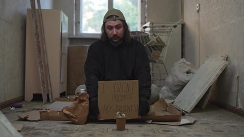 Homeless Poor Man Sitting in a Room of an Abandoned Building He is Holding a Piece of Cardboard 'I