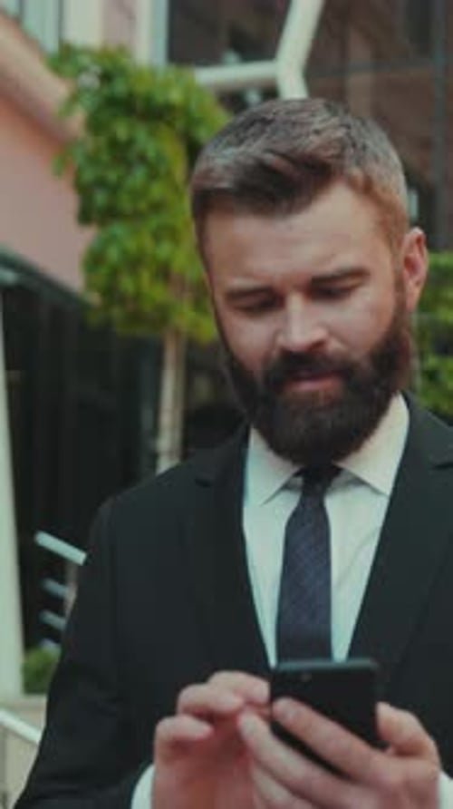Serious Successful Business Man Uses Smartphone While Walking on the Big City Business District