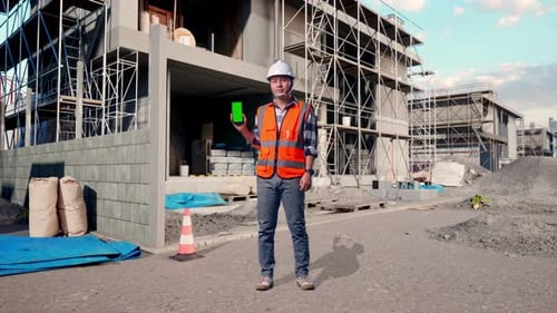 Full Body Of Male Engineer Smiling And Showing Green Screen Smartphone At Building Site