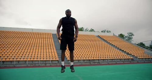 Un homme noir en tenue de sport noire faisant un exercice de burpee dans un stade avec des tribunes jaunes