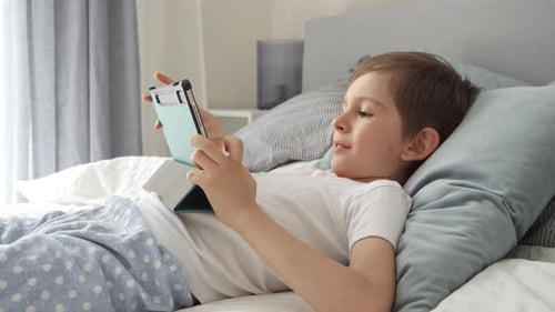 A cheerful young boy is playing with a tablet computer while lying in bed during the day