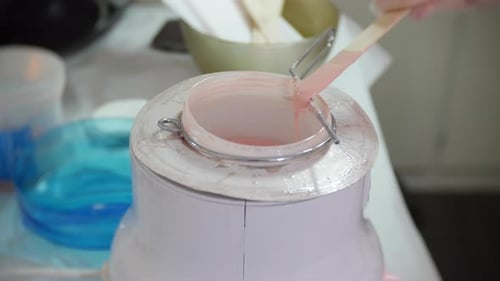 Cose up of a beautician preparing for leg waxing by applying wax to a spatula from a wax pot. The wo