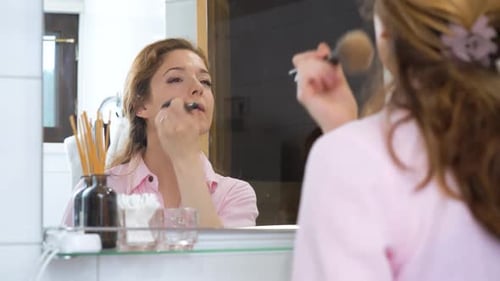 Woman Applies Makeup with Brush in Bathroom Mirror