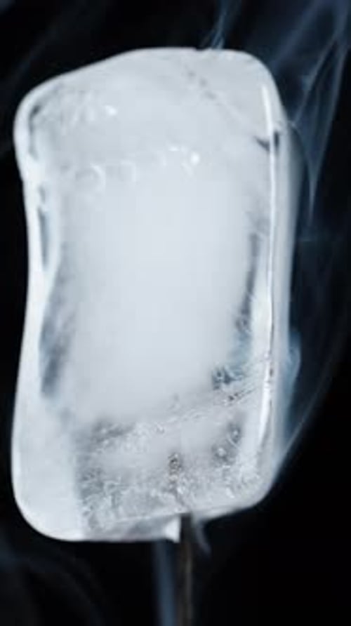 Vertical video. Melting ice cube with smoke on a black background, close-up.