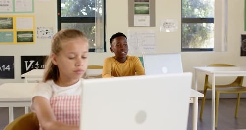 Diverse teenage classmates, boy responding to prompt, calling as girl typing on laptop in classroom