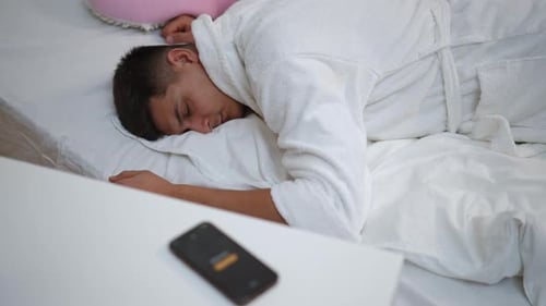 A Young Guy in a White Bathrobe Woke Up Lying on the Bed in His Bedroom From the Sound of a Mobile