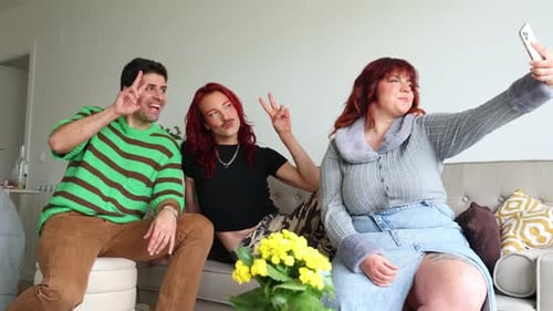 Non-binary person and plus-size friend taking selfies on sofa