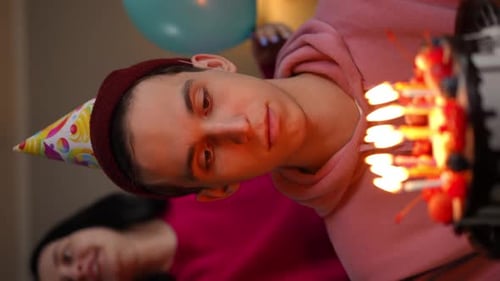 Dissatisfied Young Man with Cake with Burning Candles Indoors and Blurred Cheerful Woman Rejoicing