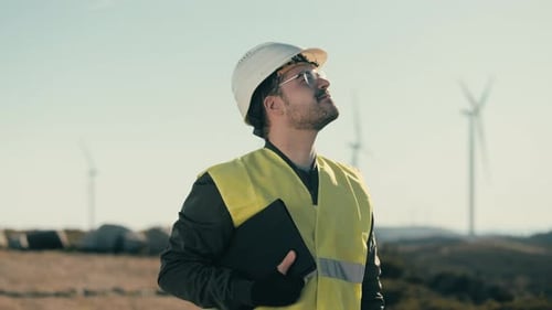A professional engineer in a field of wind turbines, dressed in safety gear, takes a break to smile