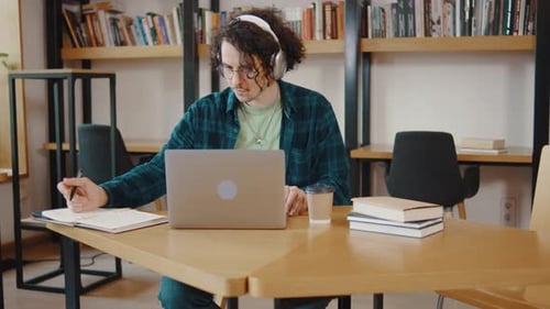 Junger Student sitzt am Schreibtisch, benutzt PC, Laptop, Computer, schaut Online-Fernunterricht