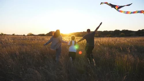 Young Parents with Small Daughter Running with Multicolored Kite Through Grass Field at Sunset