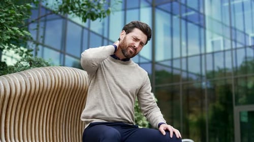 Tired businessman suffering from neck pain while sitting on a bench on the street near business