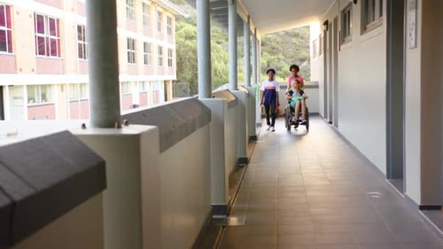Pushing wheelchair, two girl students helping friend in school hallway, smiling together