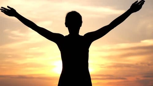 Silhouette of woman raises arms at sunset