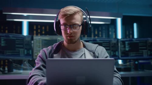 Portrait of Software Developer / Hacker / Gamer Wearing Glasses and Headset Sitting at His Desk and