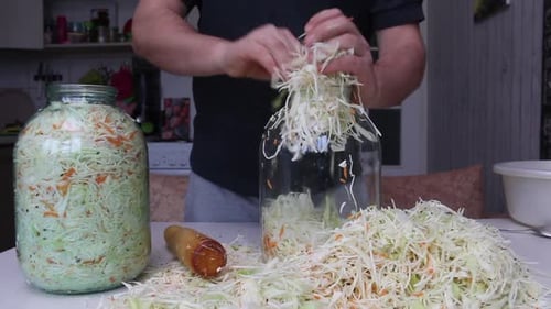 A Man Puts Chopped Cabbage And Carrots With Dill Seeds In A Jar. Compacts The Mass With A Wooden Rol