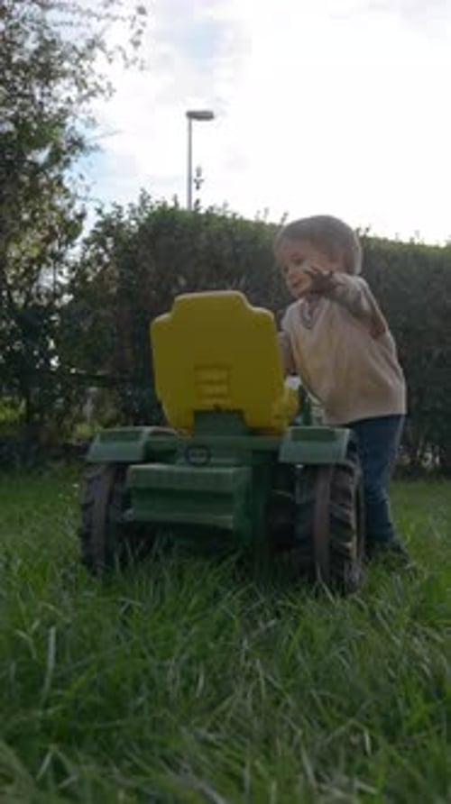 Vertical of Sweet Toddler Runs on Green Lawn to Sit on Tractor Toy