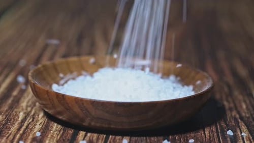 A rustic wooden bowl filled with coarse sea salt as more salt is being poured in. The salt crystals
