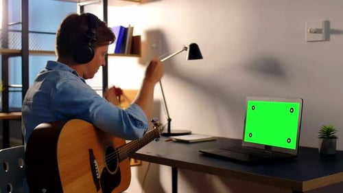 Young musician plays acoustic guitar at home with green screen laptop computer