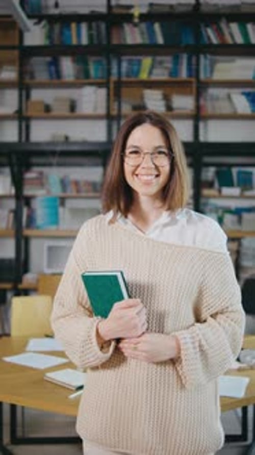 A Young Woman with Glasses in a Library Smiling Holding Book and Browsing Shelves Woman Holding Book