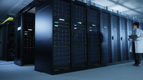 In Data Center: Male IT Specialist Wearing White Coat Walks Alongside Row of Server Racks, Uses Lap